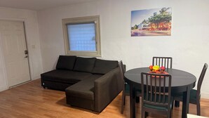 A living room with a sectional sofa, a dining table with chairs, a window with blinds, and a painting on the wall.