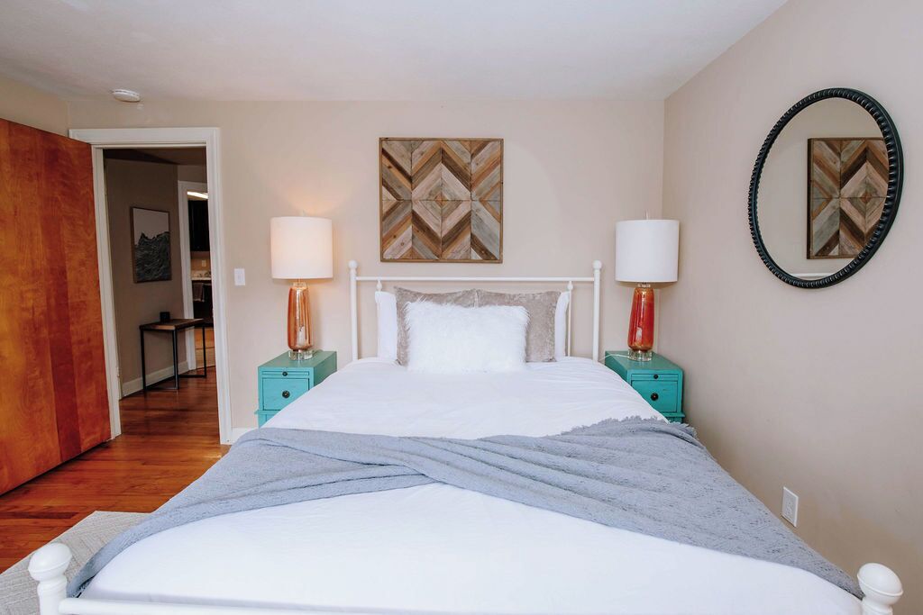 Un dormitorio con una cama blanca, dos mesitas de noche con lámparas, un espejo y una puerta que da a otra habitación.