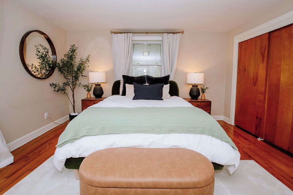 A bedroom with a bed, bedside tables, a mirror, a window with curtains, a plant, and a wardrobe.