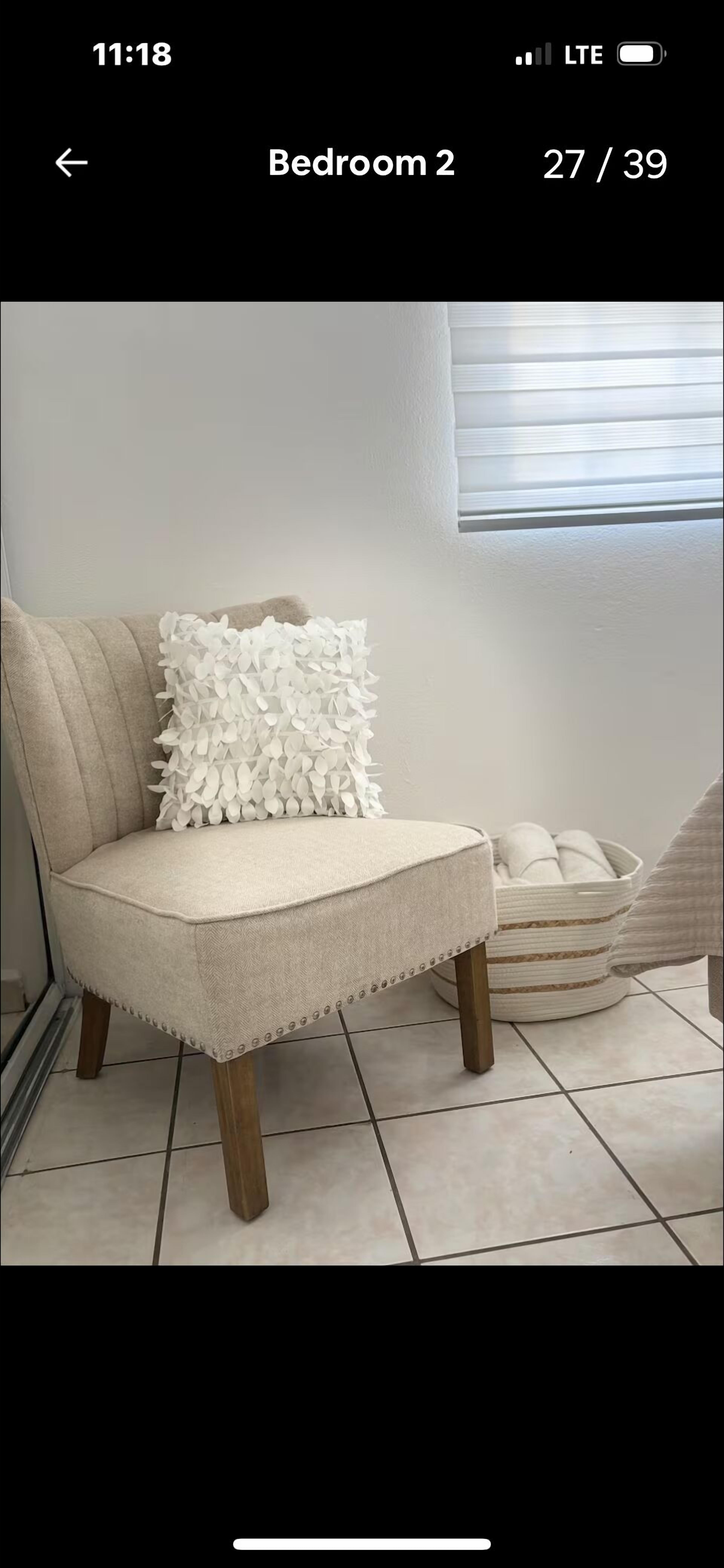 Uma poltrona bege com almofada branca texturizada, posicionada ao lado de uma cesta branca trançada em um chão de azulejos.