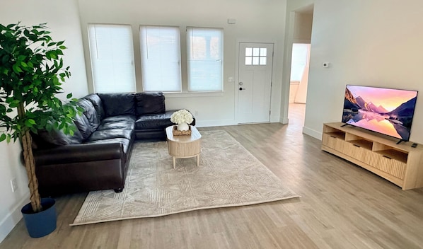 Un salon comprenant un canapé d’angle, une table basse, un téléviseur posé sur un meuble en bois et une grande fenêtre avec des stores.
