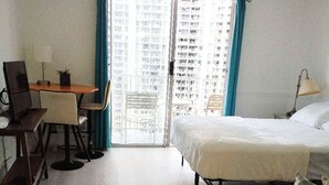 A hotel room with a bed, a desk with a computer, a chair, and a view of a high-rise building through a glass door. - In this cozy condominium located in Waikiki (Honolulu)