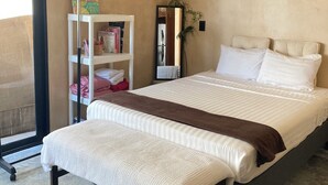 Un letto rifatto con una coperta bianco e marrone, un comodino e una mensola bianca con libri e asciugamani.