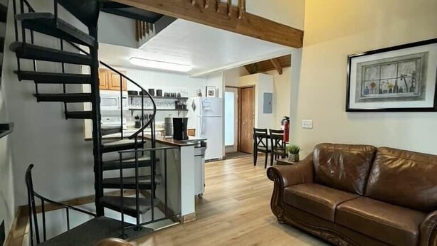 Un vaste espace de vie avec un canapé en cuir marron, un escalier en bois et une cuisine à l’arrière-plan.