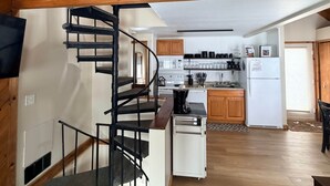 Un appartement de style « loft » avec un escalier en bois, une cuisine équipée d’appareils électroménagers blancs et un ventilateur de plafond.