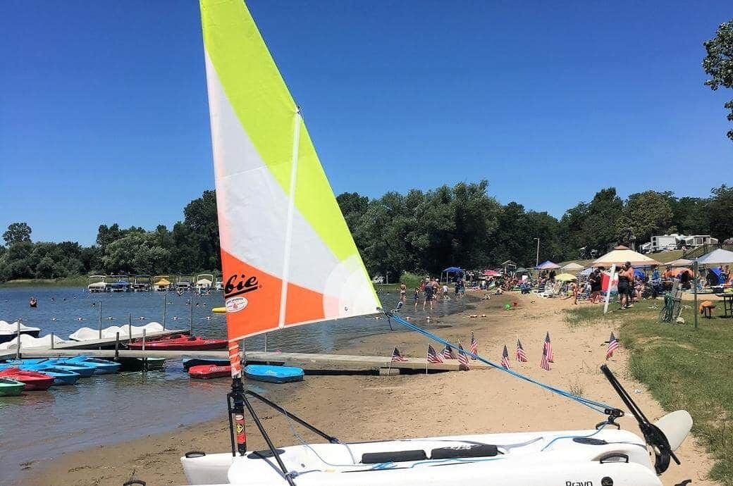 A beach with a sailboat, a dock with small boats, and a crowd of people.