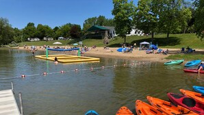 A lakeside setting with kayaks docked at a pier, a sandy beach area with people, and a building in the background.