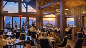 En restaurant med flere spiseborde og stole, store vinduer med udsigt til bjergene, og et udgangsskilt over døren.