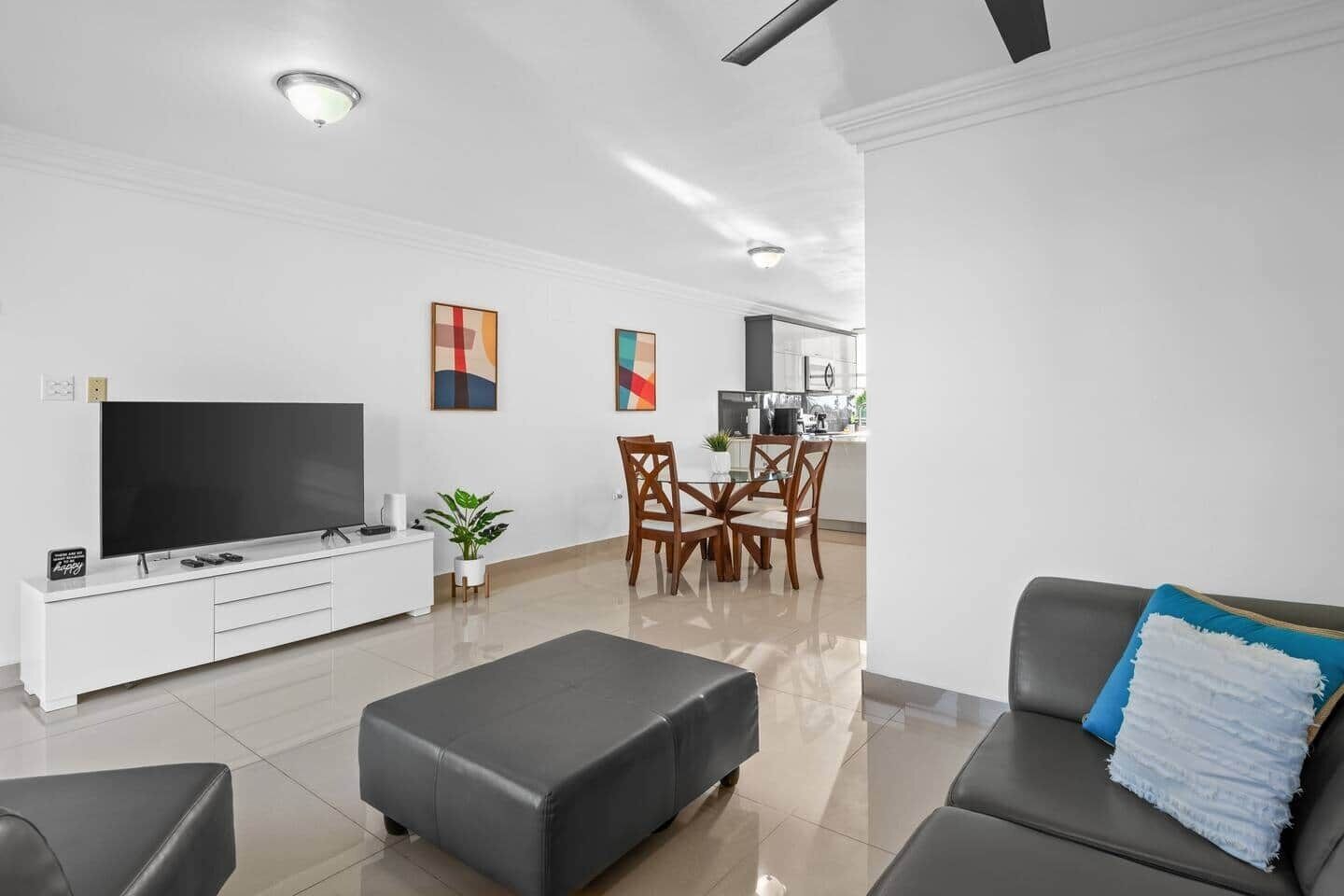 A modern living room with a flat-screen TV, a grey sofa, a dining area with wooden chairs, and a kitchen in the background.