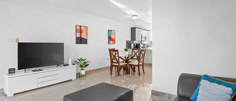 A modern living room with a flat-screen TV, a grey sofa, a dining area with wooden chairs, and a kitchen in the background.