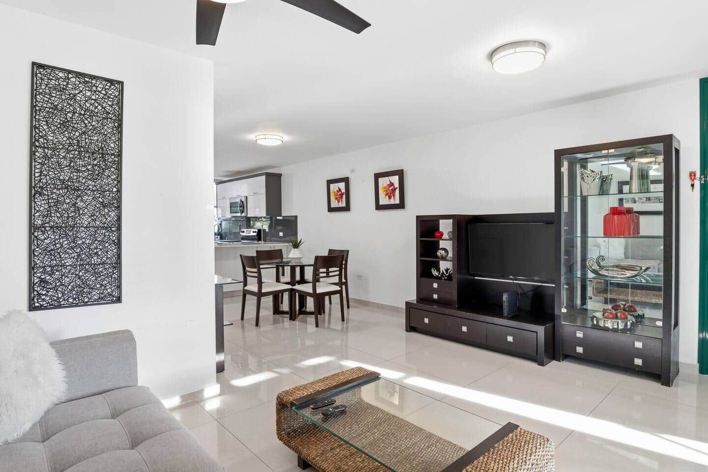A modern living room with a sofa, a glass coffee table, a TV, and a kitchen in the background.
