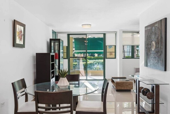 A modern dining area with a glass table, dark wooden chairs, and a view of greenery through a sliding glass door. - The Best Apartment in Aquatika (Vieques)