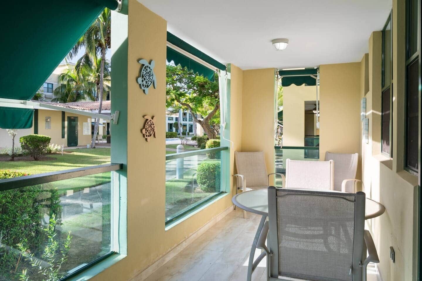A balcony with a glass railing, a table with chairs, and a view of a garden and buildings.