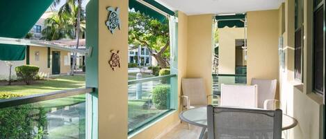 A balcony with a glass railing, a table with chairs, and a view of a garden and buildings.