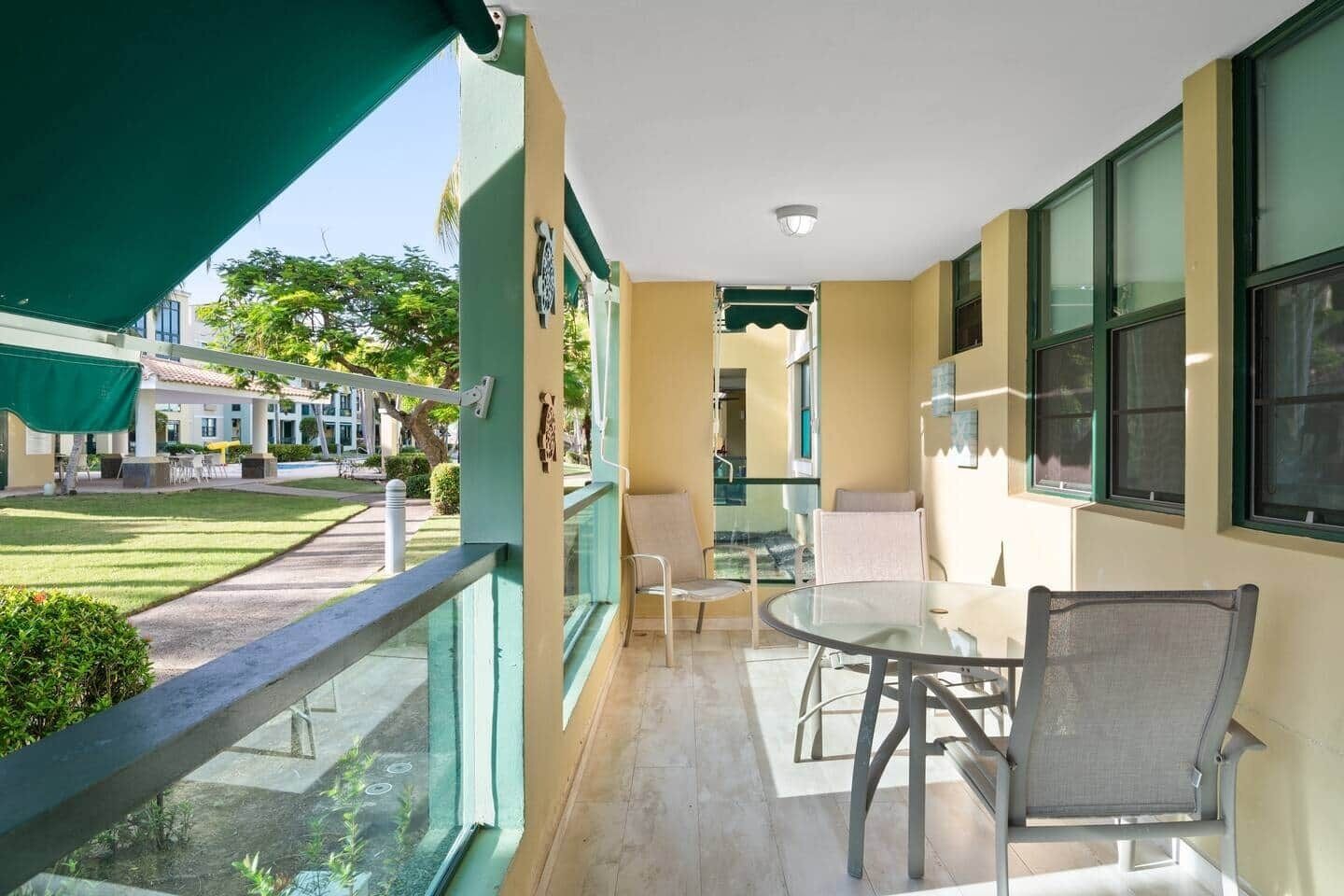 Un balcón con una mesa y sillas de vidrio, vista al patio interior y un edificio con toldo verde.