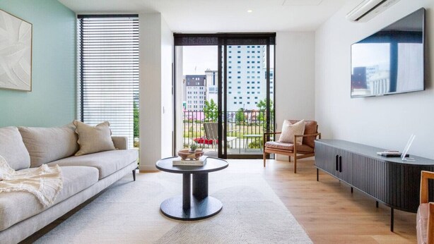 A modern living room with a sofa, a round coffee table, a chair, a TV mounted on the wall, and a view of the city through large windows. - 2-Bedroom w/ Carpark. Overlooks Central East Frame (Christchurch)