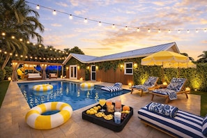 Un área de piscina con sillones de descanso, una mesa con aperitivos y luces colgantes.