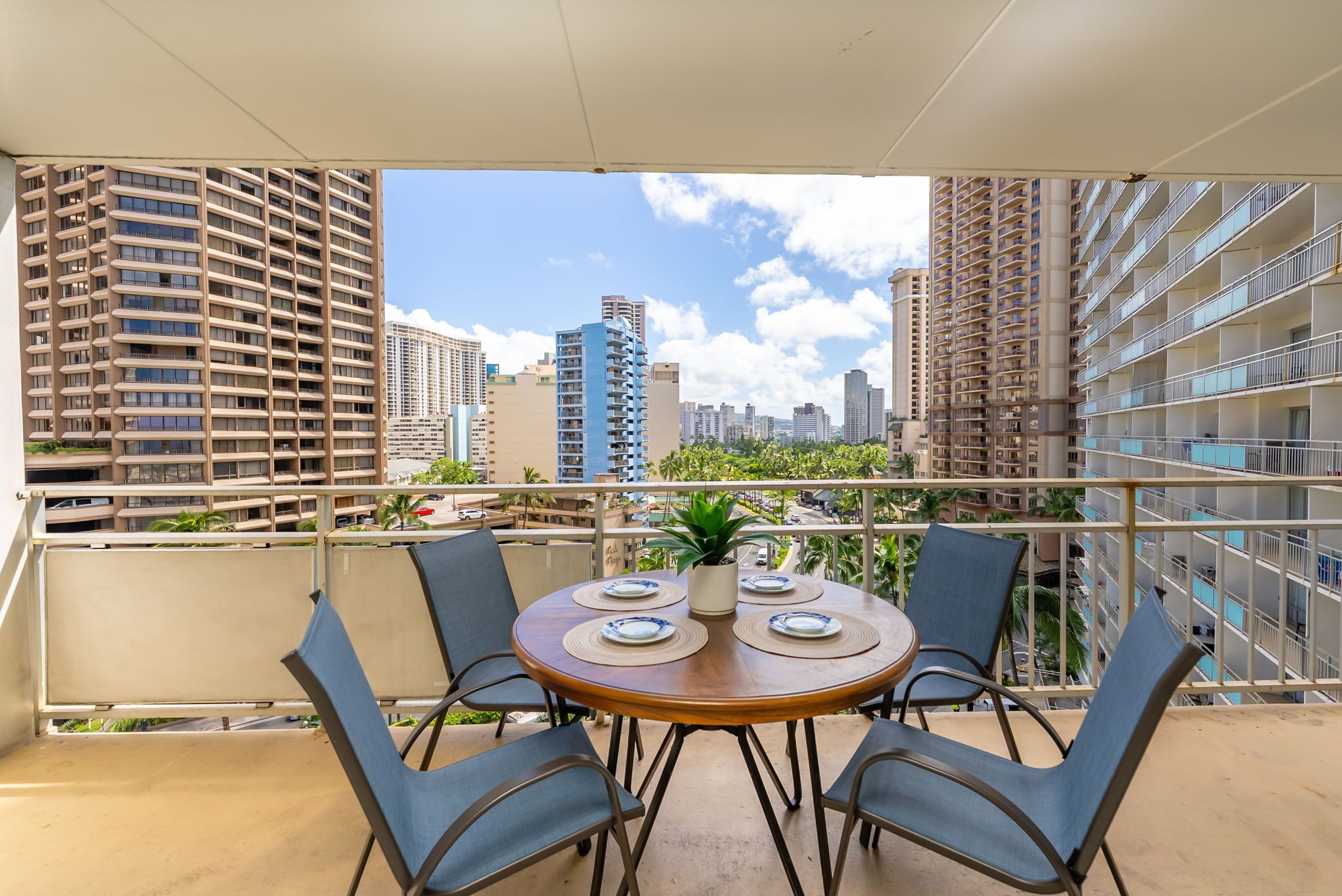 A balcony with a round table and four chairs, overlooking a cityscape with tall buildings and a clear sky.