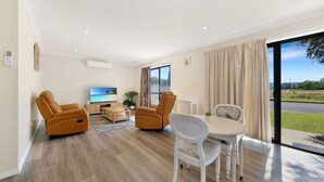 A modern living room with a flat-screen TV, two leather recliners, a wooden coffee table, and a dining table with white chairs.