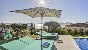 Et poolområde med to grønne liggestole, en stor parasol og udsigt til havet og bakker.