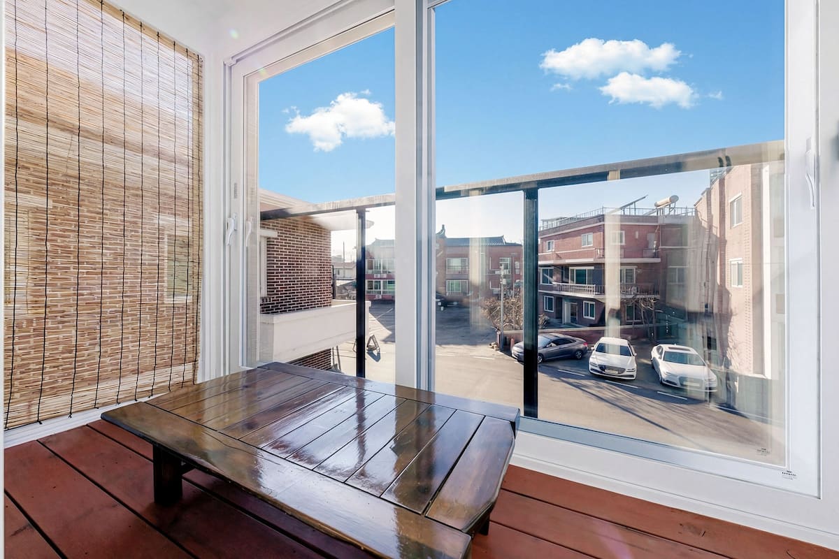 Un balcon avec un banc en bois, de grandes fenêtres et une vue sur une rue bordée d’immeubles et de voitures garées.