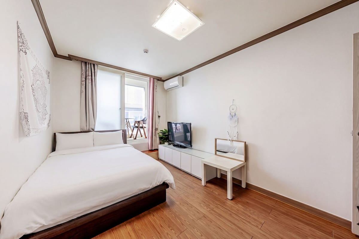 Une chambre à coucher avec un lit, un bureau, une télévision et une fenêtre avec des rideaux.