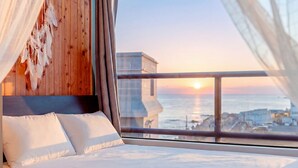 Une chambre à coucher avec une tête de lit en bois, du linge de lit blanc et une vue sur le coucher de soleil sur l’océan.