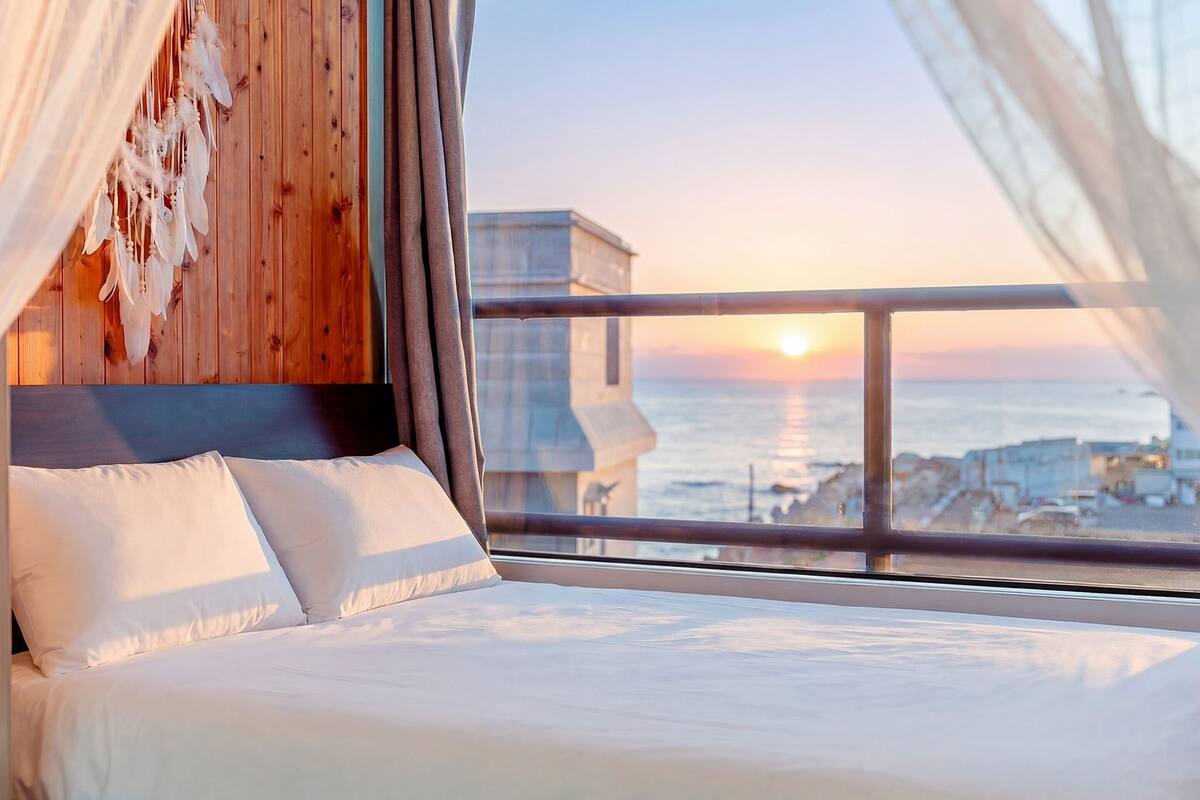 Une chambre à coucher avec une tête de lit en bois, du linge de lit blanc et une vue sur le coucher de soleil sur l’océan.