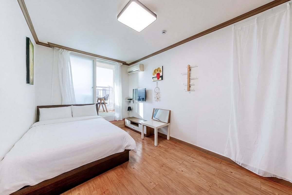 Une chambre d’hôtel avec un plancher en bois, un lit avec une literie blanche, un bureau avec une chaise, une télévision et une fenêtre avec des rideaux.