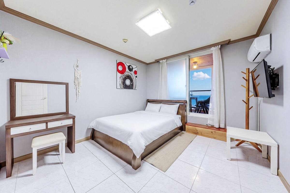 Une chambre à coucher comprenant un lit, un bureau, un banc, un téléviseur fixé au mur et une vue sur l’extérieur.