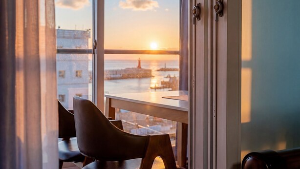 Une vue depuis une fenêtre donnant sur un coucher de soleil au-dessus d’une étendue d’eau, avec une chaise et une table à l’intérieur d’une pièce.