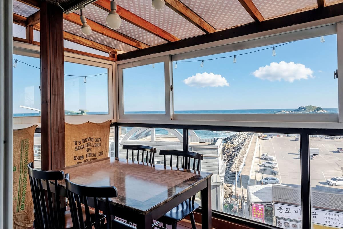 Un espace repas avec une grande table et des chaises, de grandes fenêtres donnant sur la mer et un parking.