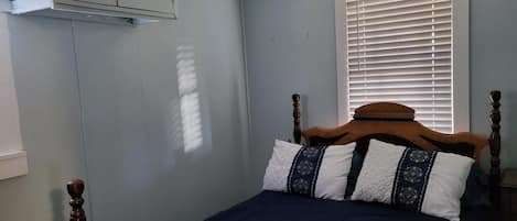 A bedroom with a wooden bed frame, a dark blue bedspread, and patterned pillows. There is a window with blinds, a white wall, and a white ceiling.