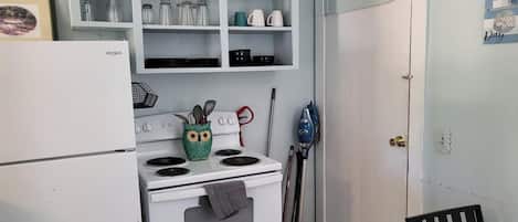 A compact kitchen with a white stove, a green owl-shaped pot holder, and a blue vacuum cleaner.