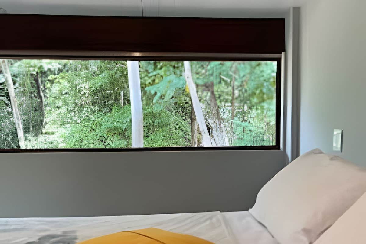 A bed with pillows, a window with a forest view, and a white ceiling.