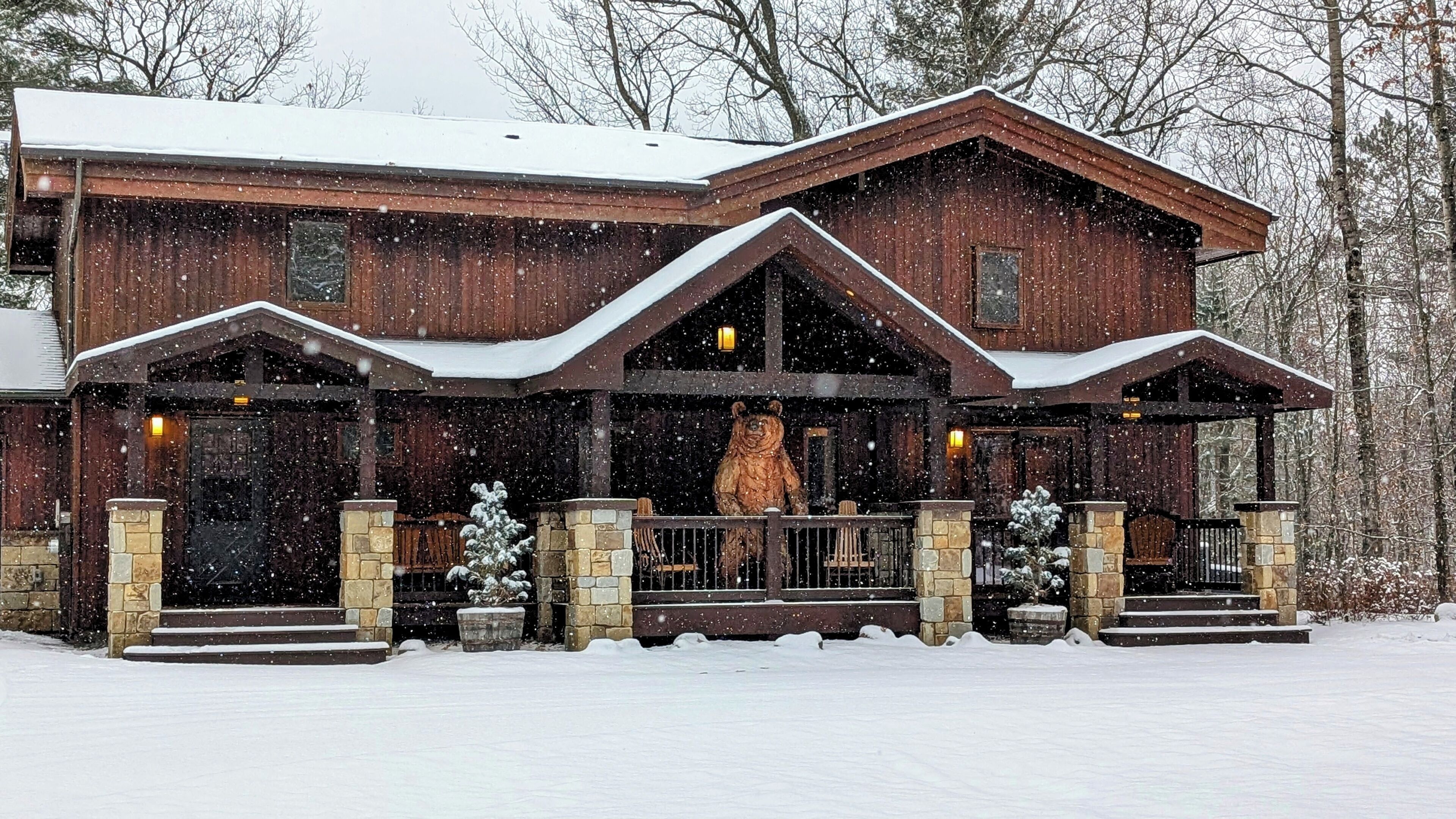 Une grande cabane en bois, dont le toit est recouvert de neige et qui possède plusieurs cheminées, est entourée d’un paysage enneigé.