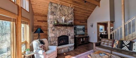 Ein gemütliches Wohnzimmer mit Kamin aus Stein, Holzboden und Blick ins Freie.