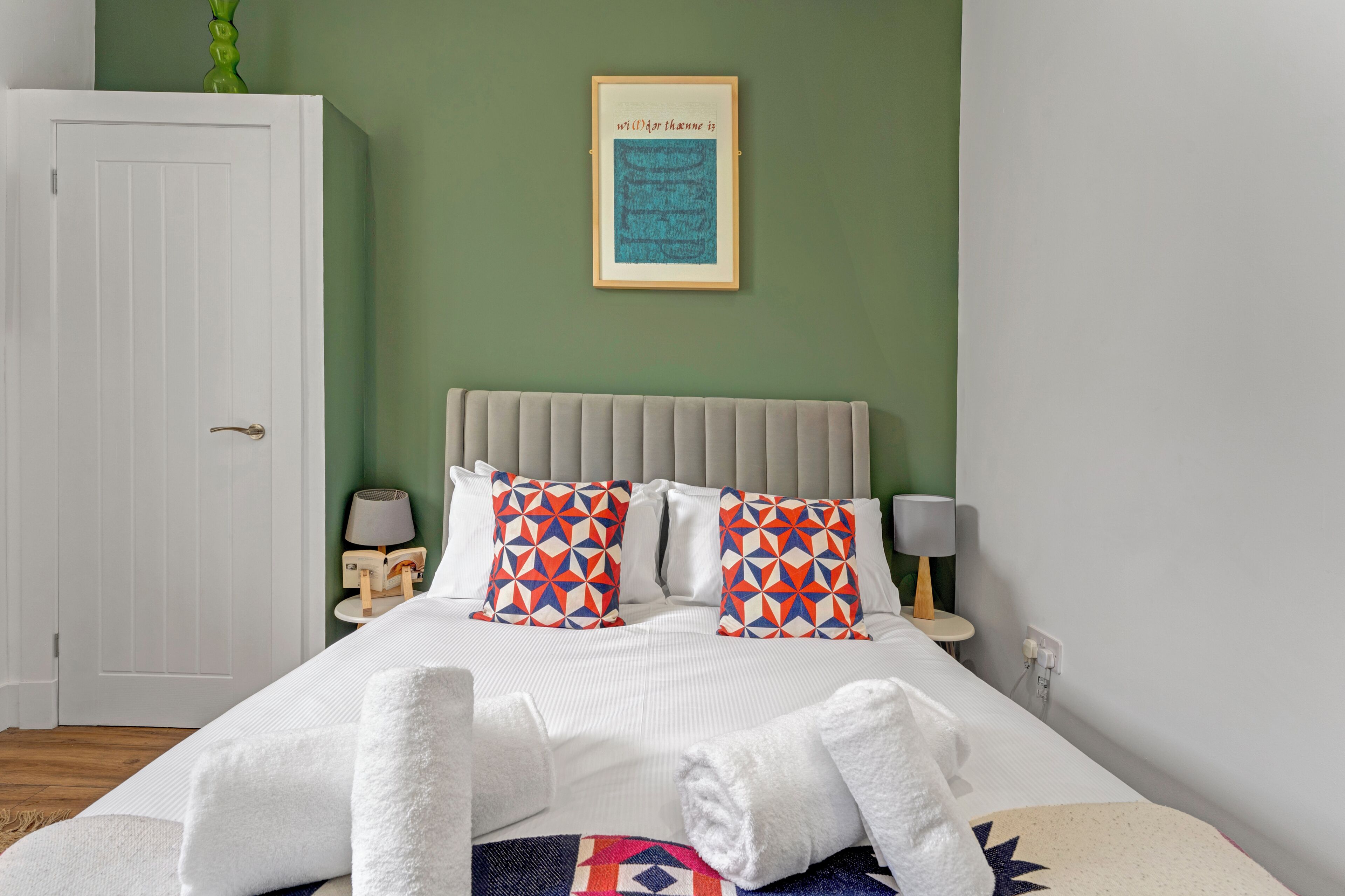Cama bem arrumada com lençóis brancos e almofadas estampadas, mesa de cabeceira com abajur e uma frase emoldurada na parede.