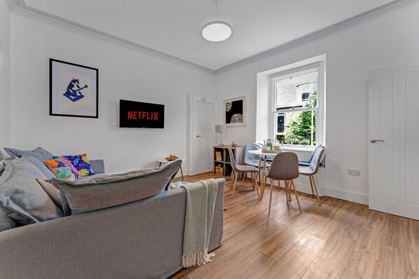Una sala moderna con un sofá gris, un comedor con mesa y sillas, una estantería y un ventanal con vistas a zonas verdes.
