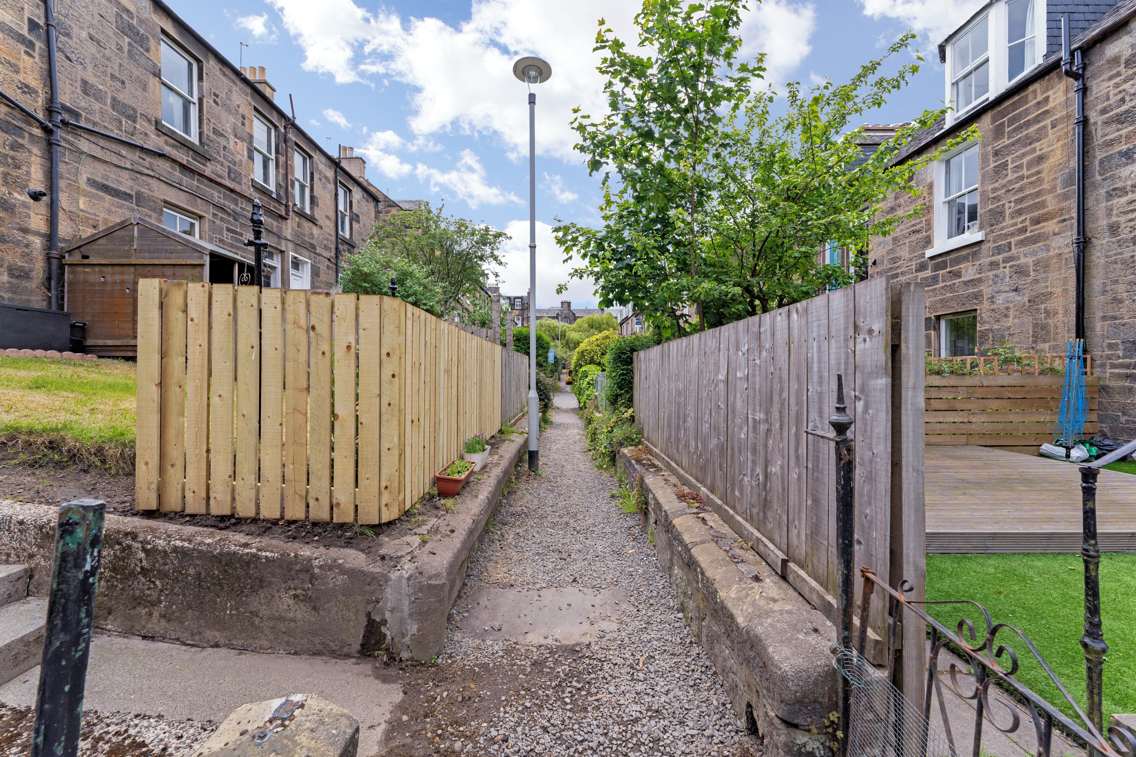 A narrow alleyway between two stone buildings with a wooden fence on one side and a metal fence on the other.