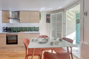 A modern kitchen with wooden cabinets, a built-in oven, and a dining area with a white table and orange chairs. - Appartement Aurelia - Welkeys (Bandol)