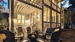 Un espace extérieur aménagé, abrité et doté de fauteuils Adirondack et d’un foyer, attenant à une maison avec de grandes fenêtres et une véranda.