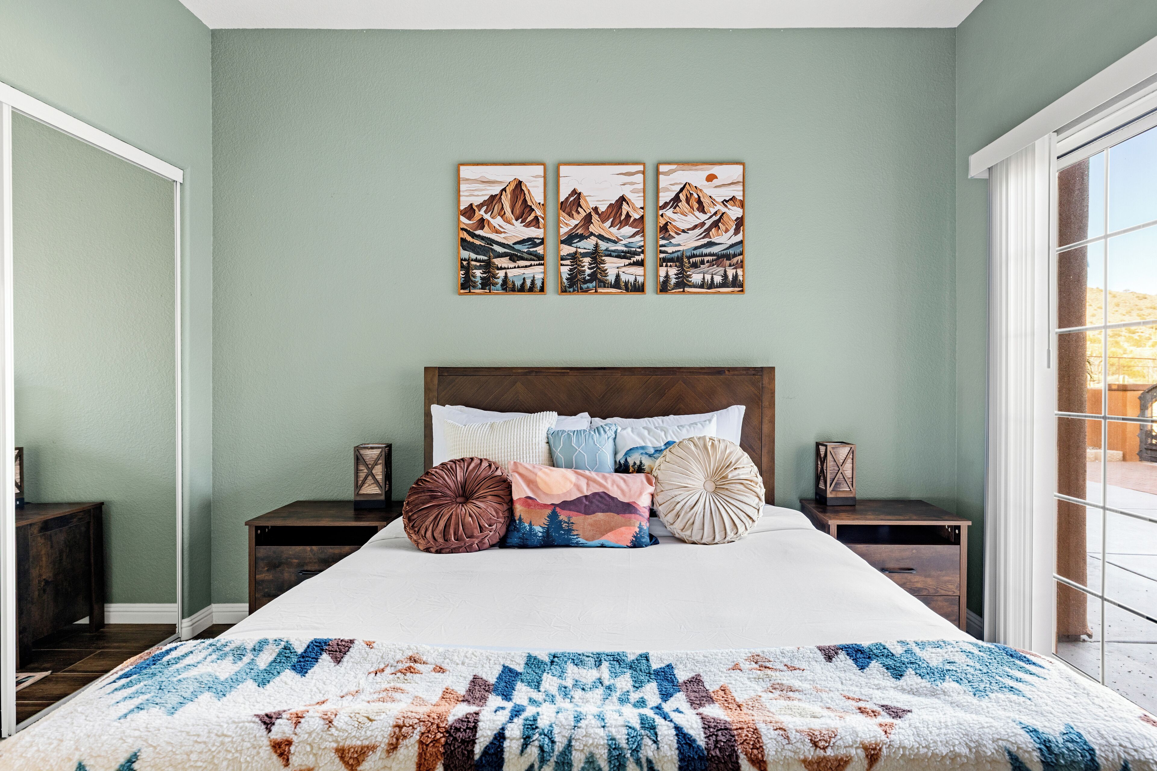 Un dormitorio con una cama grande, varios almohadones, un cuadro en la pared y vistas a través de un ventanal corredero.