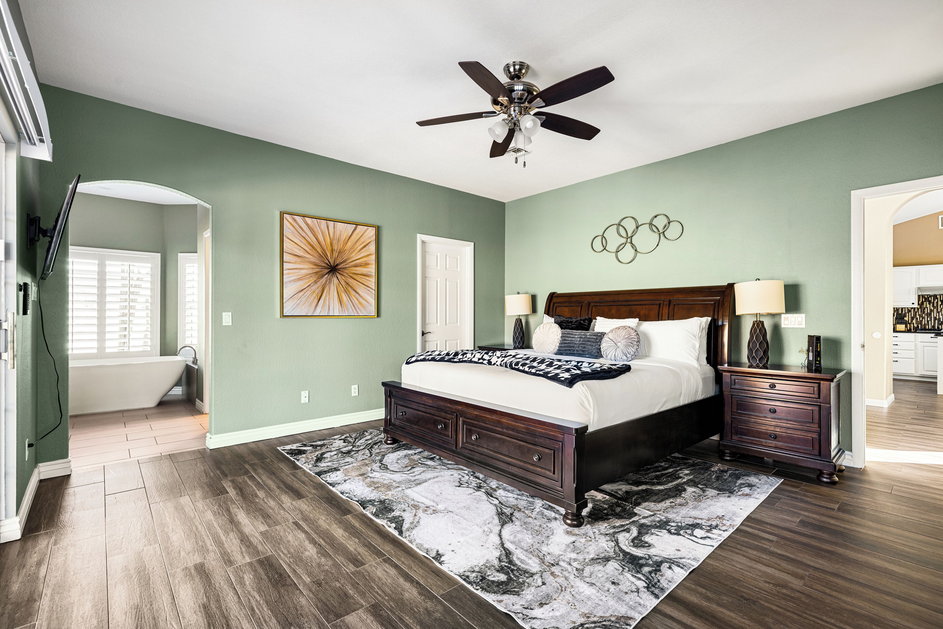 A bedroom with a bed, nightstands, a ceiling fan, a large abstract painting, and a view of the bathroom.