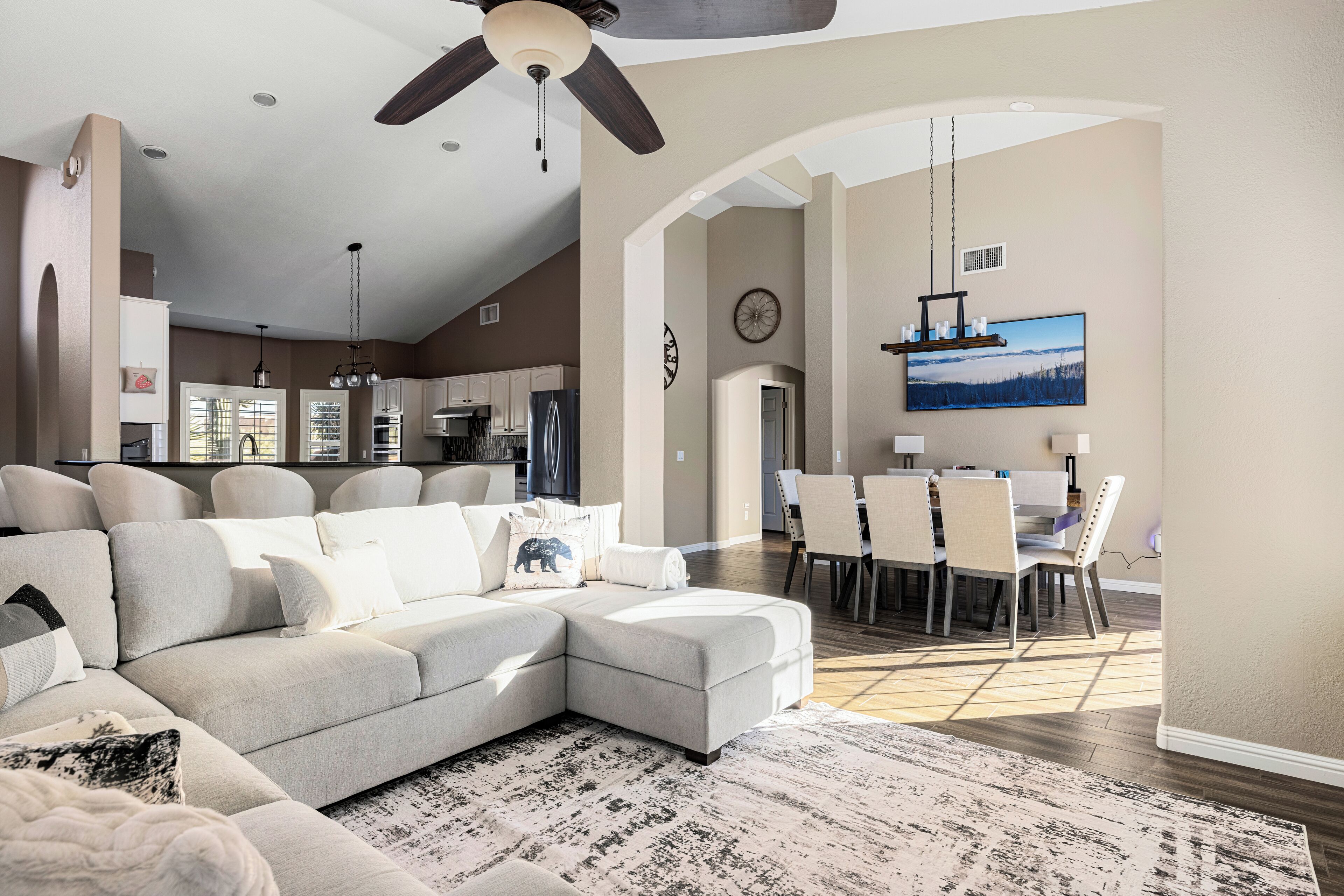 A spacious living room with a sectional sofa, a dining area with a table and chairs, a kitchen in the background, and a ceiling fan.