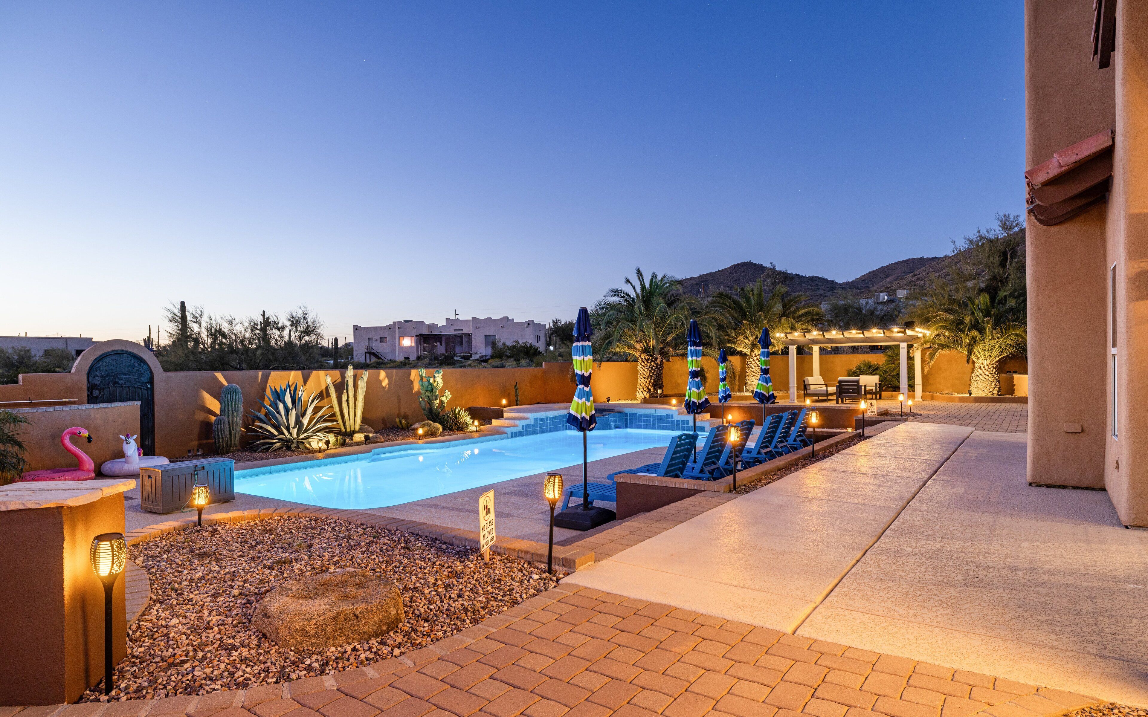 A swimming pool with lounge chairs and umbrellas, surrounded by desert plants and a building in the background.