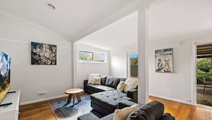 Un salon moderne avec un canapé gris, une table basse ronde en bois et un tapis coloré. Deux fenêtres laissent entrer la lumière naturelle et offrent une vue sur la verdure à l’extérieur.