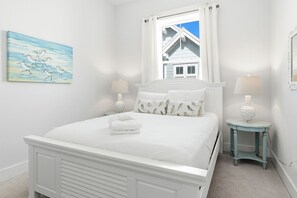 A white bedroom with a bed, two bedside tables, a window with curtains, a painting on the wall, and a view of a house outside. - Power & wifi on - 3 BR, close to the resort style pool, Big Chill & Bikes (Santa Rosa Beach)