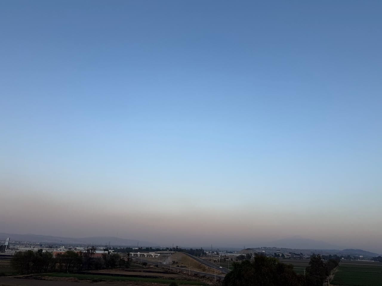 A wide view of a landscape with a clear sky, distant buildings, and open fields.