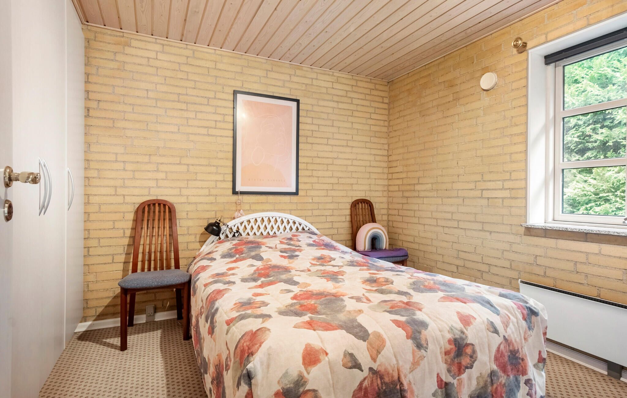 A bedroom with a brick wall, a bed with a floral bedspread, a wooden chair, a small table with a lamp, and a window with a view of trees.